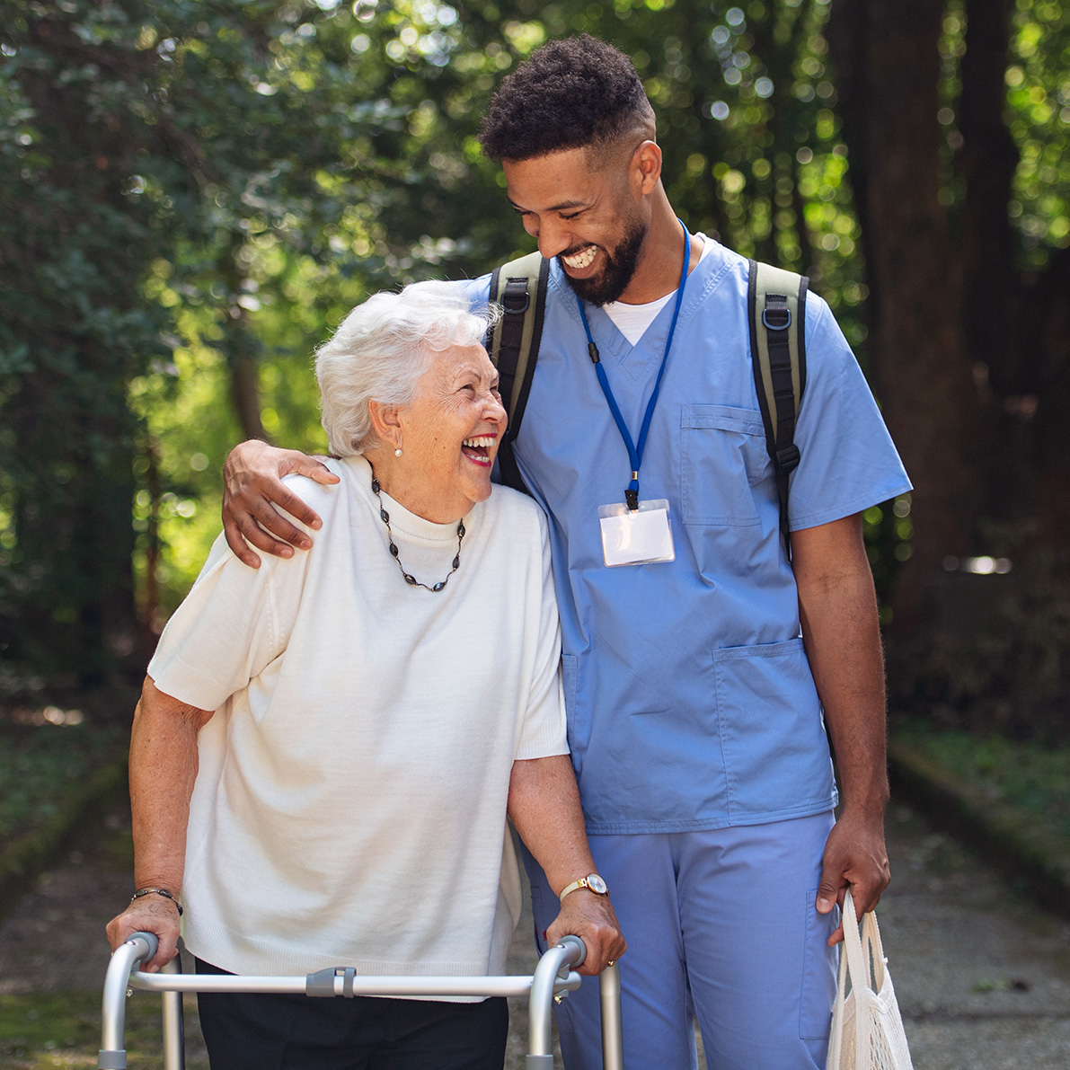 Healthcare worker in scrubs assists an elderly person with a walker on a tree-lined path.